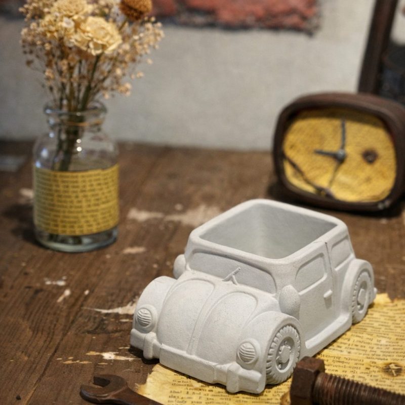 ヴィンテージ風の木製テーブルの上に置かれたセメント製クラシックカー型オブジェ。ドライフラワーとアンティーク時計と組み合わせた無機質インテリア。